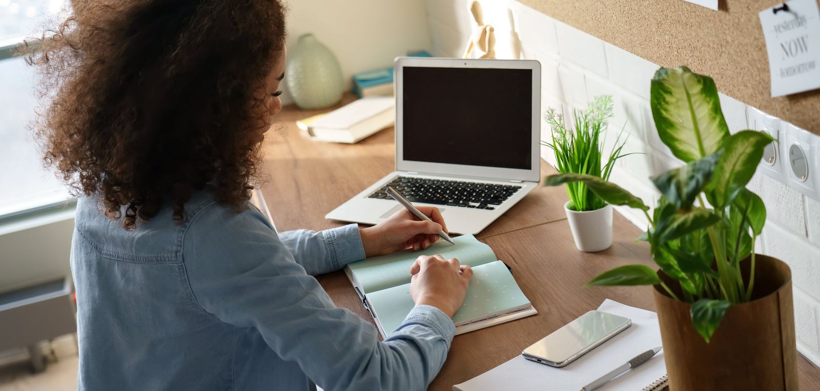 Designer writing in a notebook at a sunlit desk beside a laptop and plants